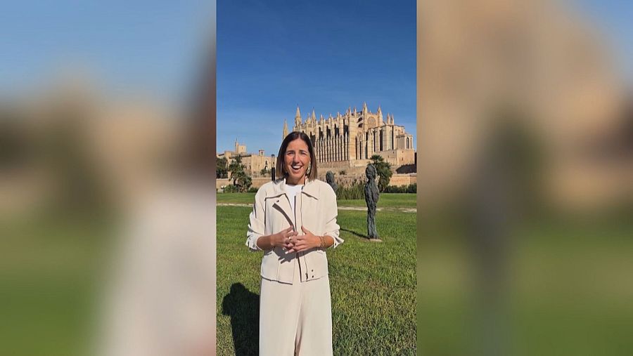 Una reportera sonriente en traje beige se encuentra en un parque urbano frente a la fachada de una catedral gótica bajo un cielo azul.