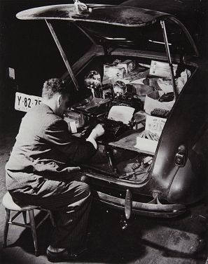 Weegee sentado en un taburete escribiendo a maquina en el maletero de su Chevrolet