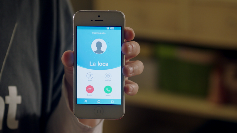 En la imatge apareix una mà subjectant un telèfon mòbil blanc la pantalla del qual està il·luminada ja que està rebent una trucada d'un contacte guardat com 