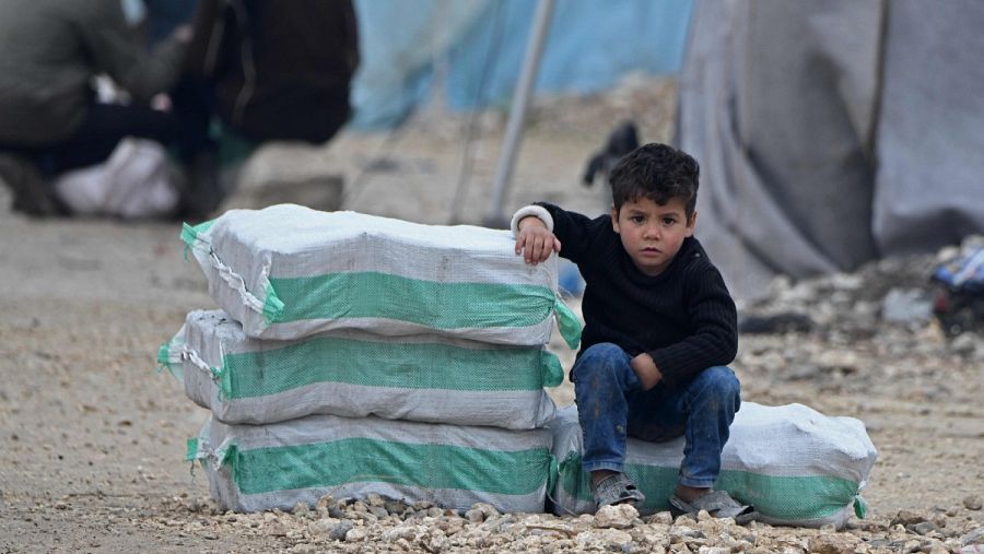 Un niño sirio desplazado sentado junto a paquetes de ayuda humanitaria