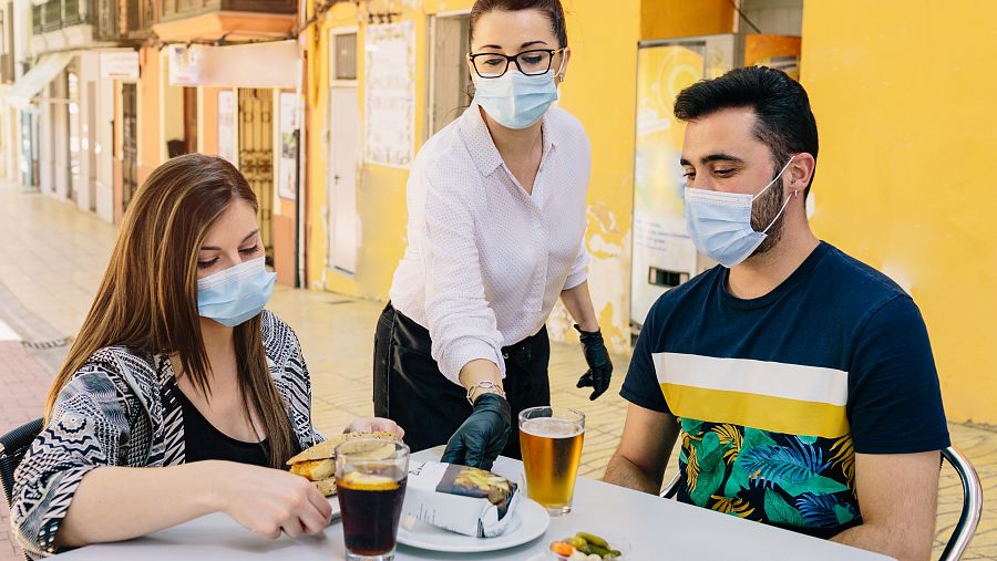 Una camarera atiende a dos clientes en la terraza de un bar con mascarilla.