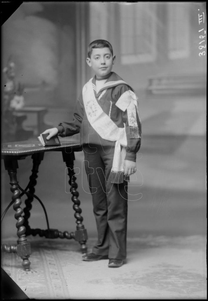 NI&Ntilde;O VESTIDO DE PRIMERA COMUNION, DE PIE JUNTO A UNA MESA