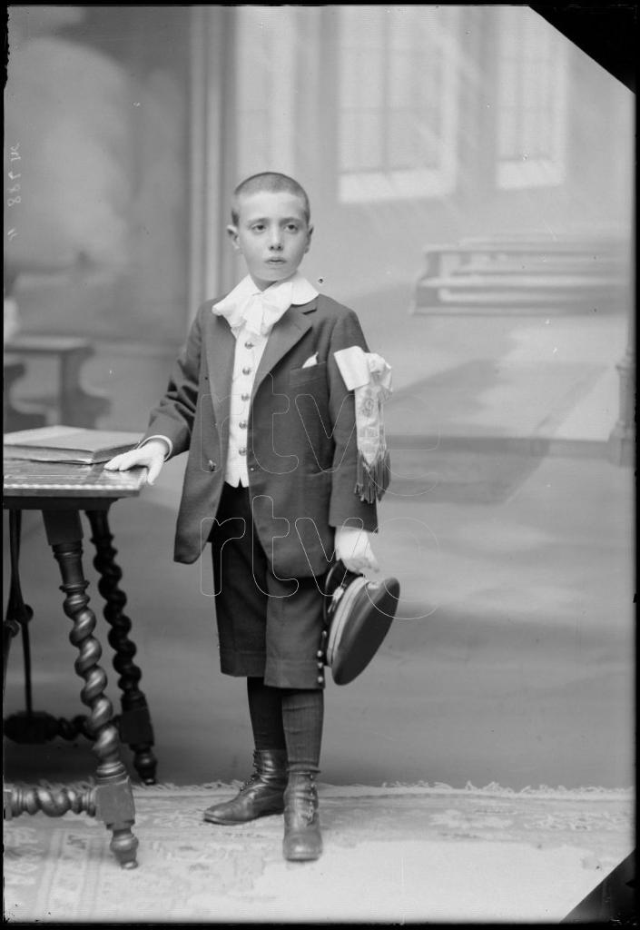 NIÑO VESTIDO DE PRIMERA COMUNION, DE PIE JUNTO A UNA MESA