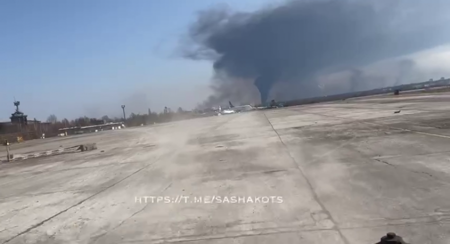 Muestra la grabación desde un vehiculo militar que abandona el aeródromo de Hostomel, con humo saliendo del fondo de la imagen.