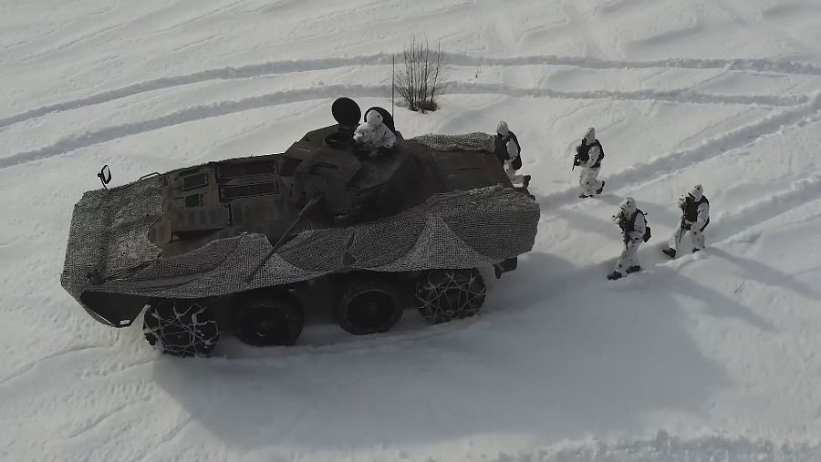 Un carro de combate rodeado de militares avanza sobre la nieve visto desde una altura.