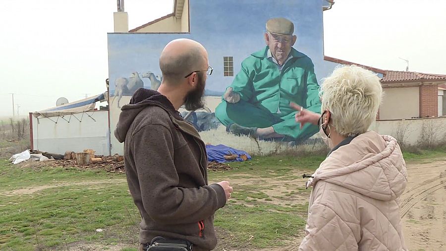 El artista explica a la reportera una de sus pinturas al aire libre en el pequeño pueblo de Nava de la Asunción.