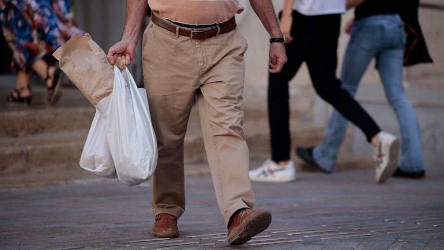 Pies de un señor caminando por la calle con dos bolsas de plástico y alimentos.