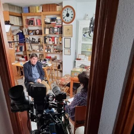 Luis García Montero rodeado de libros frente a la entrevistadora y la cámara enmarcados por la puerta abierta.