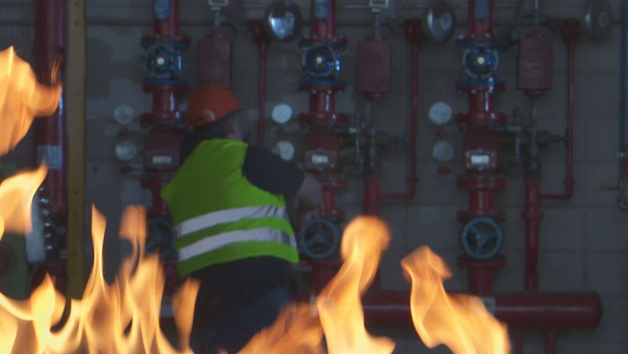 Un hombre con chaleco reflectante junto a tuberías y llaves con llamas de fuego en primer plano