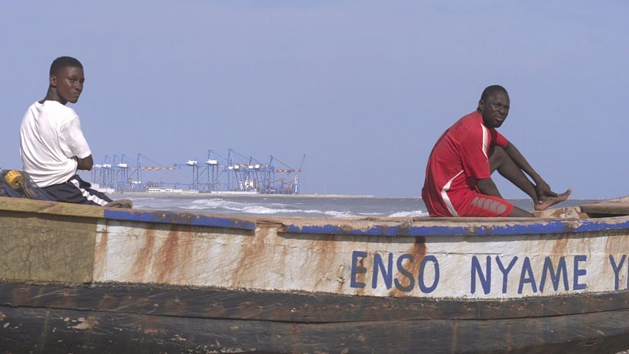 Dos jóvenes pescadores en una barcaza con las grúas del puerto al fondo.