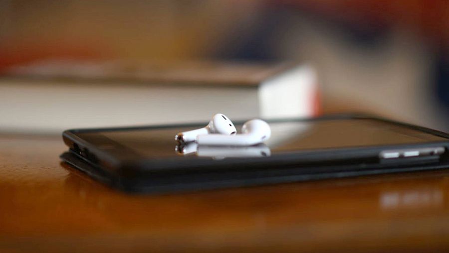 Imagen de dos auriculares inalámbricos sobre un dispositivo móvil sobre una mesa, con un libro de fondo