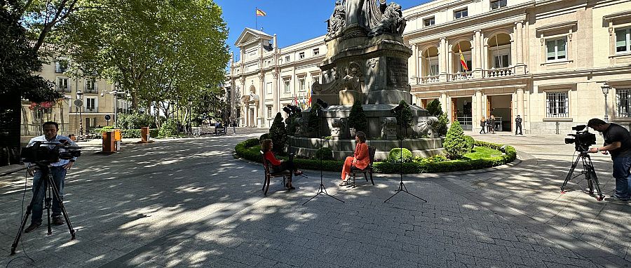 Dos cámaras de televisión en una plaza donde hay dos mujeres sentadas en sendas sillas bajo un cielo azul con edificios neoclásicos al fondo.