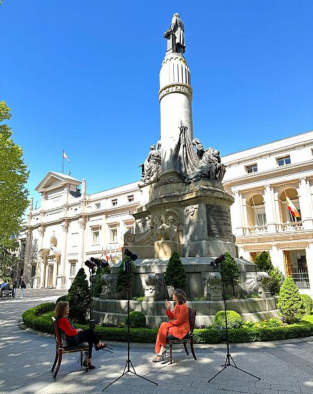 Dos mujeres sentadas en sendas sillas en mitad de una plaza con un obelisco y la fachada neoclásica del Senado al fondo con un cielo despejado y azul.