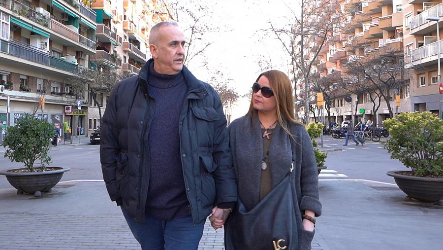 Un hombre y una mujer pasean de frente a la cámara cogidos de la mano por la calle de una ciudad.