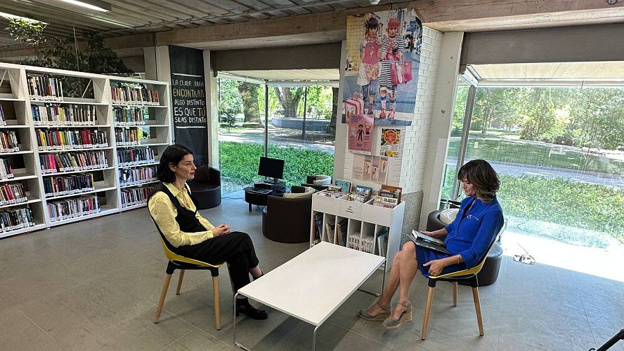 Dos mujeres conversan sentadas en sillas de madera dentro de un espacio rodeadaas de estanterías con libros y grandes ventanales.