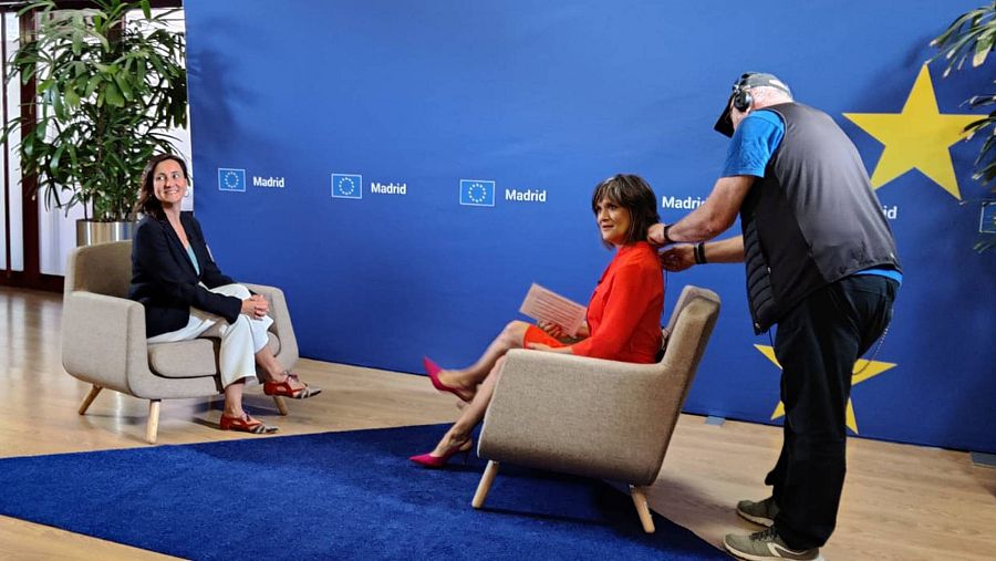 Untécnico de sonido coloca el micrófono a una mujer vestida de rojo y sentada frente a otra en sillas con la bandera azul de estrellas amarillas en el fondo.
