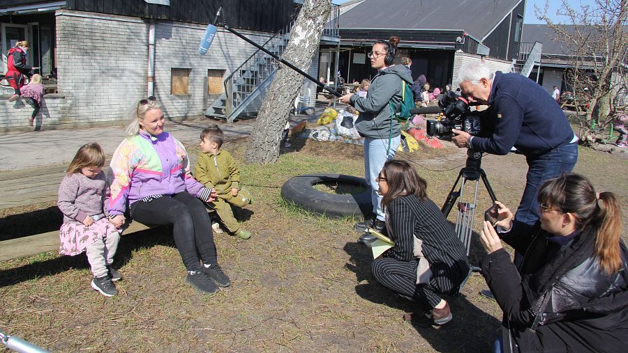 Un equipo de televisión se dispone a entrevistar a una mujer con dos niños al aire libre con otros niños en las inmediaciones.