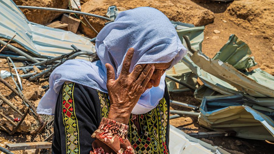 Una anciana con velo morado se tapa la cara. Tras ella se ven unas ruinas.