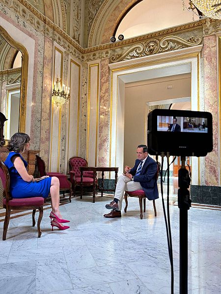 Una mujer entrevista a un hombre sentados en sillas y se les ve a través de un monitor.