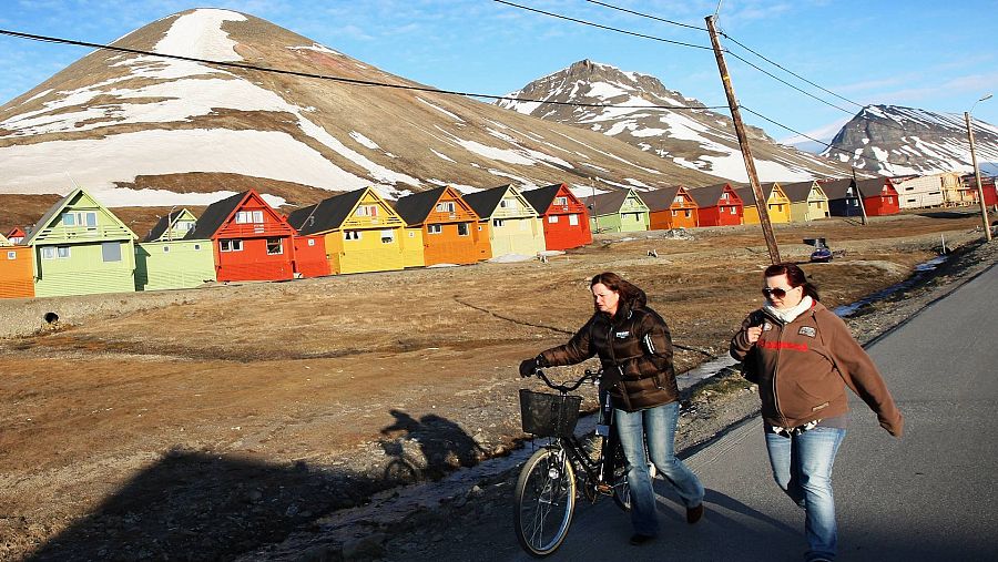 Imagen de Longyearbyen, con montañas nevadas al fondo, casas de colores y dos transeuntes en primer plano