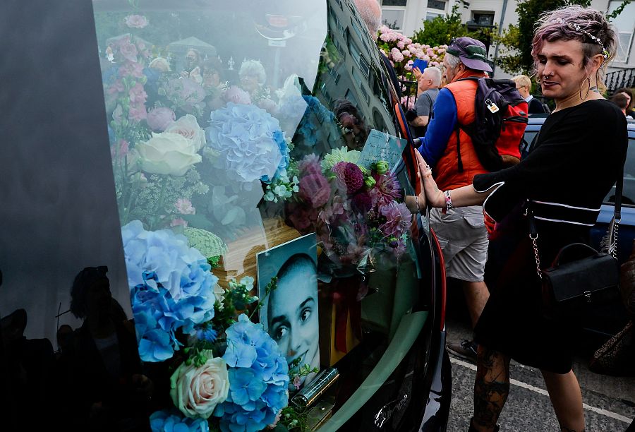 Una persona acompaña a pie el coche fúnebre, en el que se ven flores y una foto de O'Connor