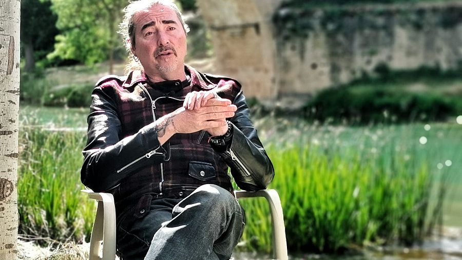 Hombre con coleta y pelo canoso, sentado y  vestido con cazadora de cuero negro con vegetación a su espalda.