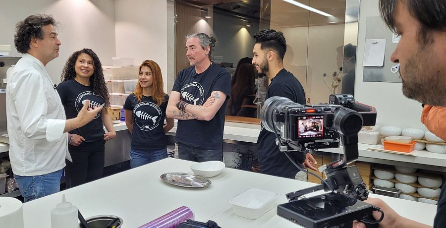 Dos cocineros con chaquetilla blanca y alumnos de hostelería con camisetas negras y logotipo de raspa de pescado en el pecho.