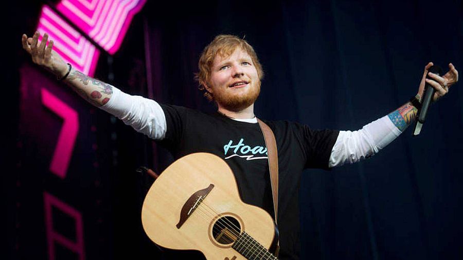 Ed Sheeran sonríe durante un concierto, con la guitarra colgada y los brazos abiertos.