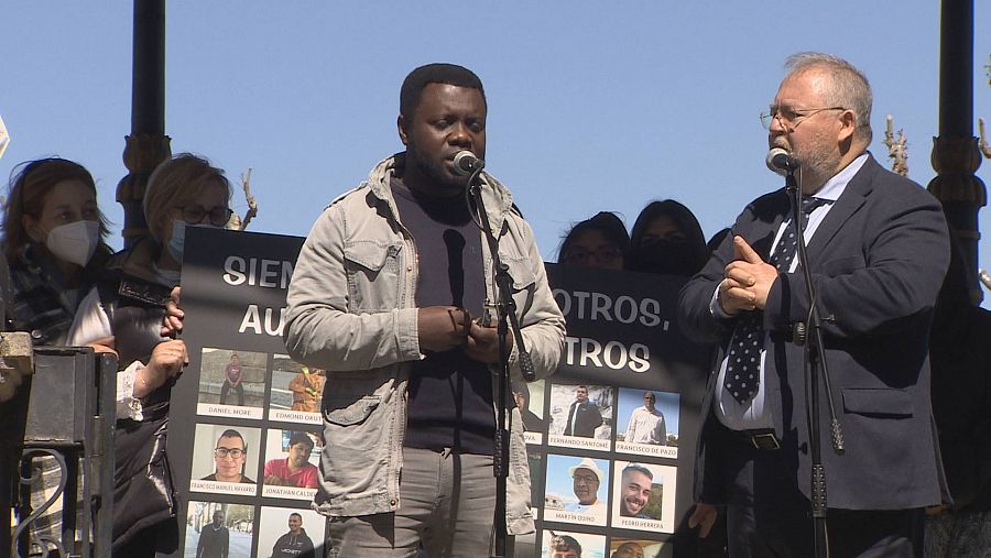 Una persona de raza negra habla en un micrófono, en la calle, flanqueada por carteles con fotos de personas.
