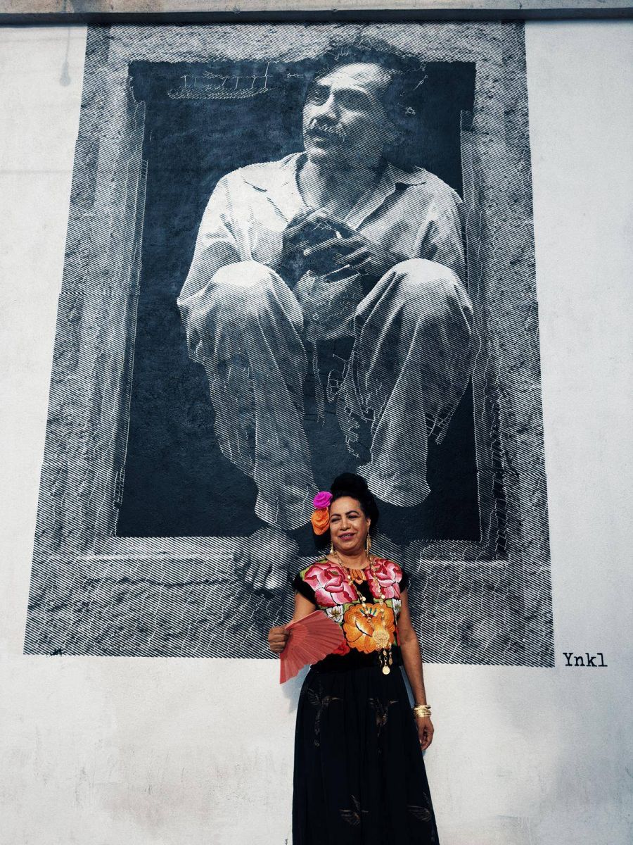 Un indígena juchiteco vestido de mujer posa delante de una pared donde hay un retrato de un señor.