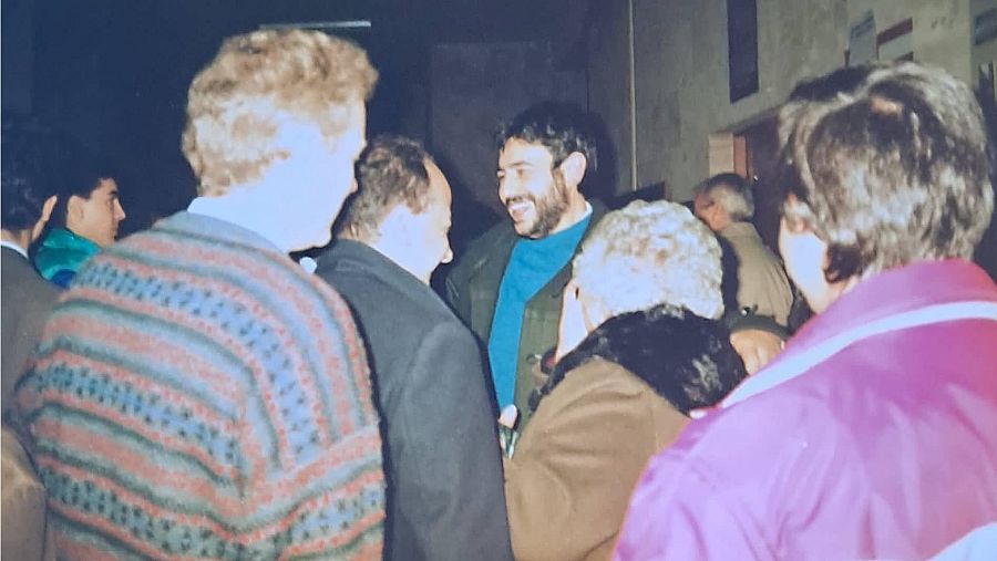 Un joven con barba rodeado de gente de espaldas que le quiere saludar.