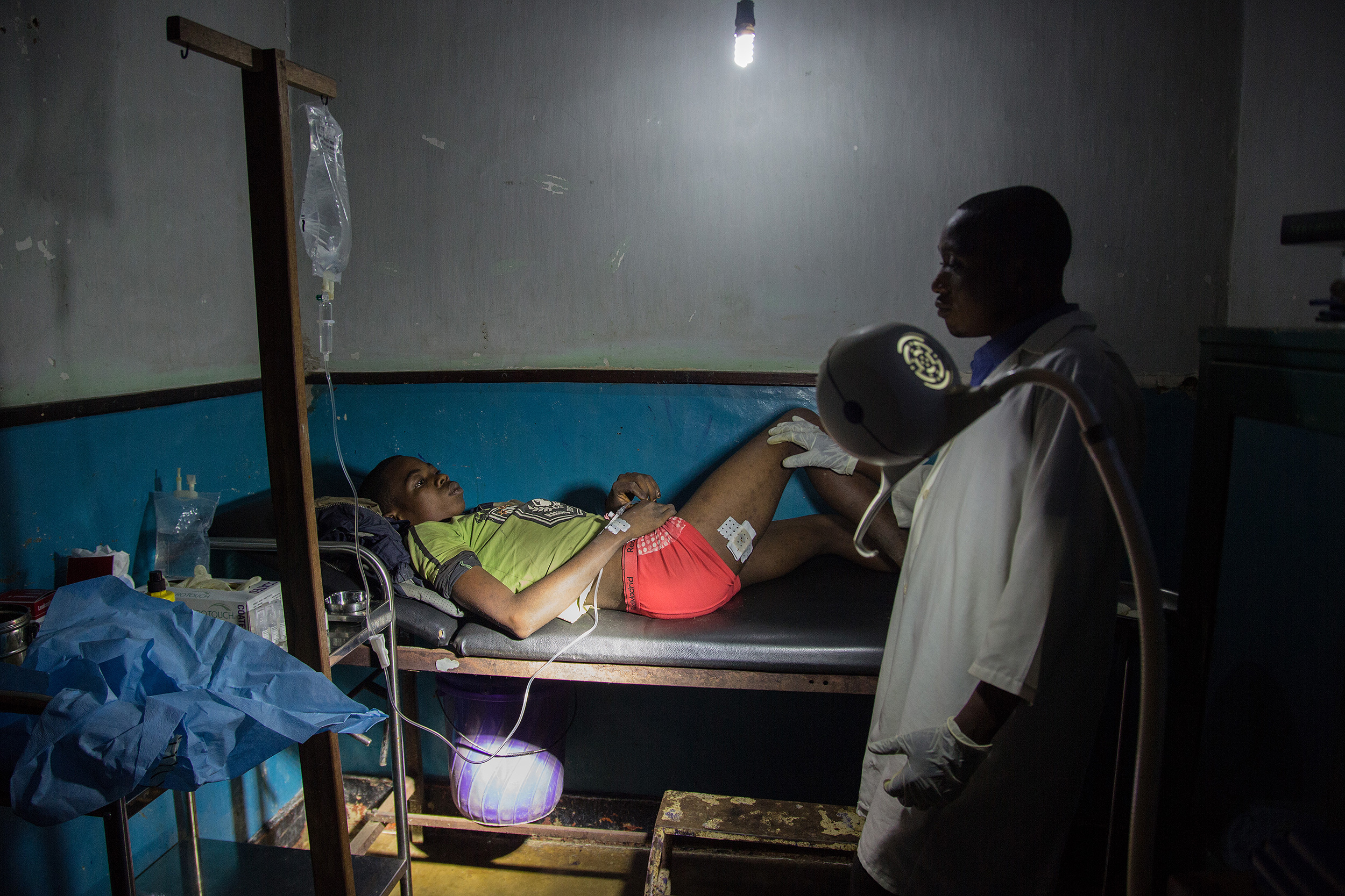 Un joven yace en una cama del hospital de Lulingu, bajo la supervisión de un médico. Luce un vendaje en una de sus piernas.