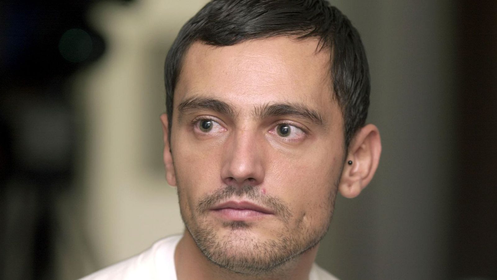 Retrato de un hombre con cabello y barba cortos, oscuro, y un pendiente; expresión seria, mirando a la izquierda; fondo oscuro y desenfocado.