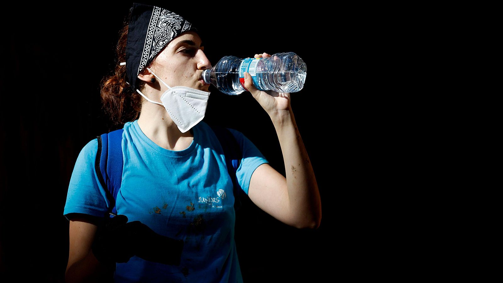 Una voluntaria con una mascarilla blanca y un pañuelo en la cabeza bebe agua de una botella durante un descanso. 
