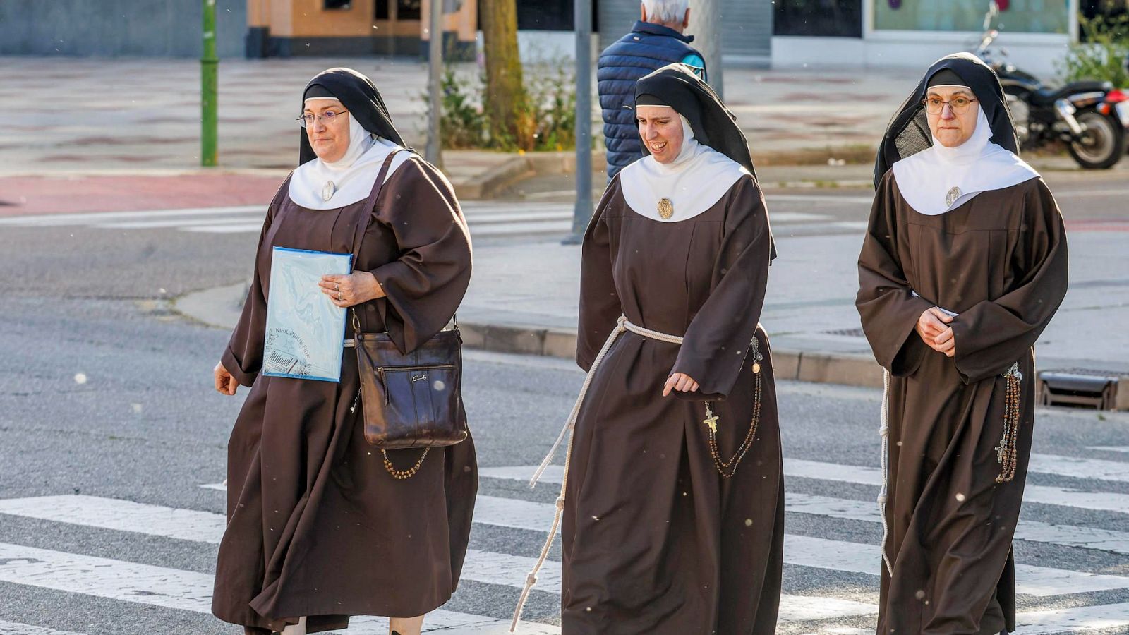 Tres monjas con hábitos marrones y rosarios cruzan la calle por un paso de peatones.