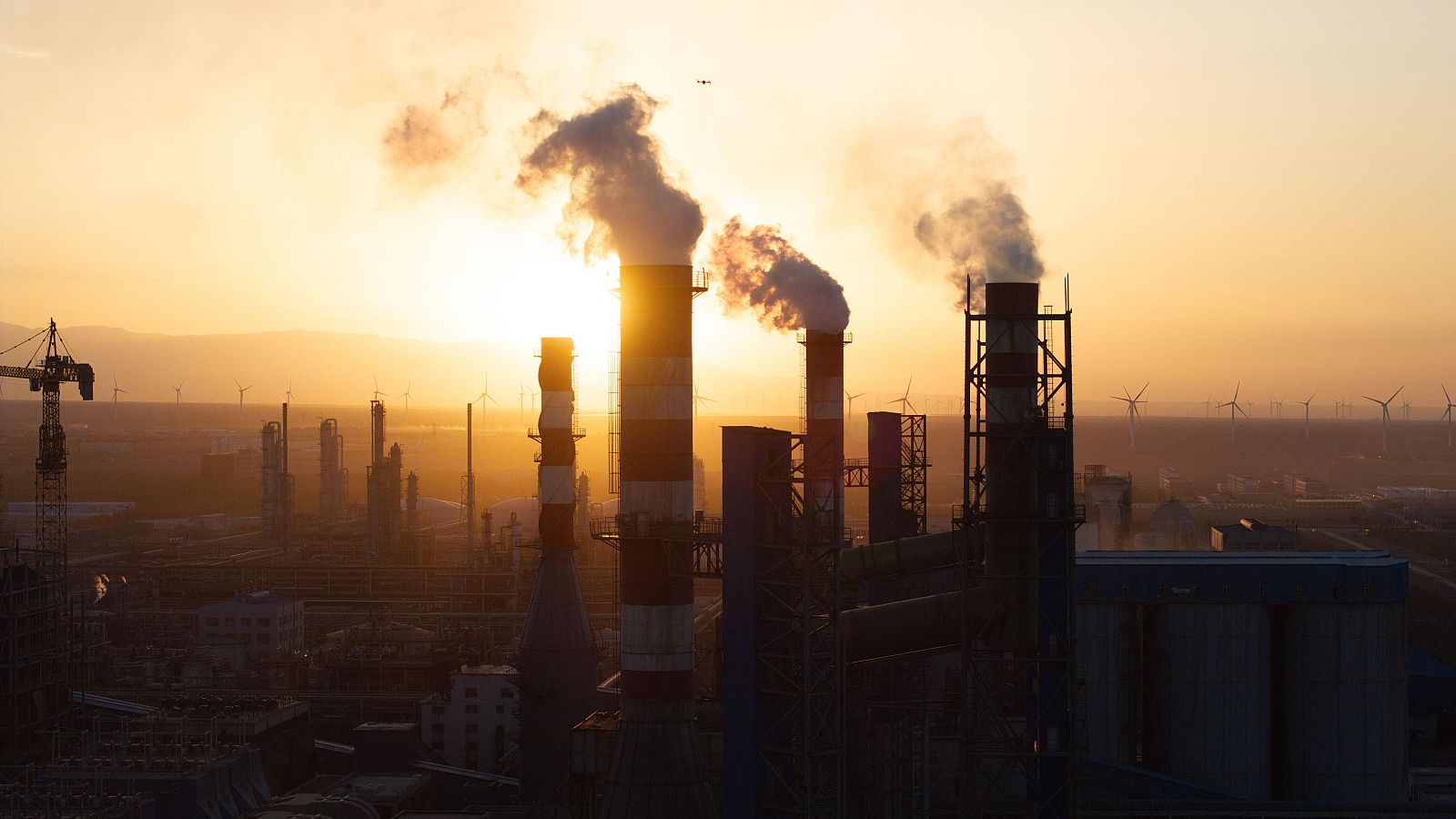 Humo sale de las chimeneas de una planta industrial al atardecer, con un parque eólico visible al fondo.