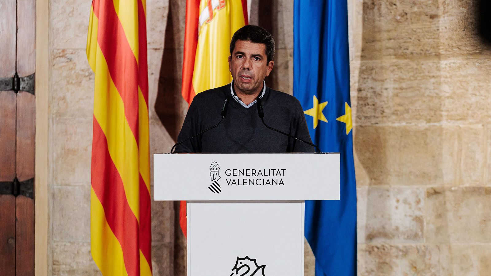 El presidente de la Generalitat Valenciana, Carlos Mazón, habla en un atril con las banderas de la Comunidad Valenciana, España y la UE detrás.