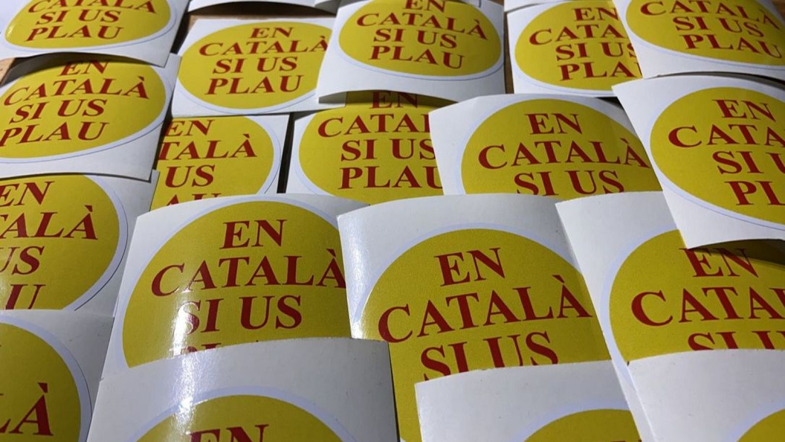 Múltiples pegatinas amarillas con letras rojas de la campaña "En Català, Si Us Plau" de Òmnium Cultural.
