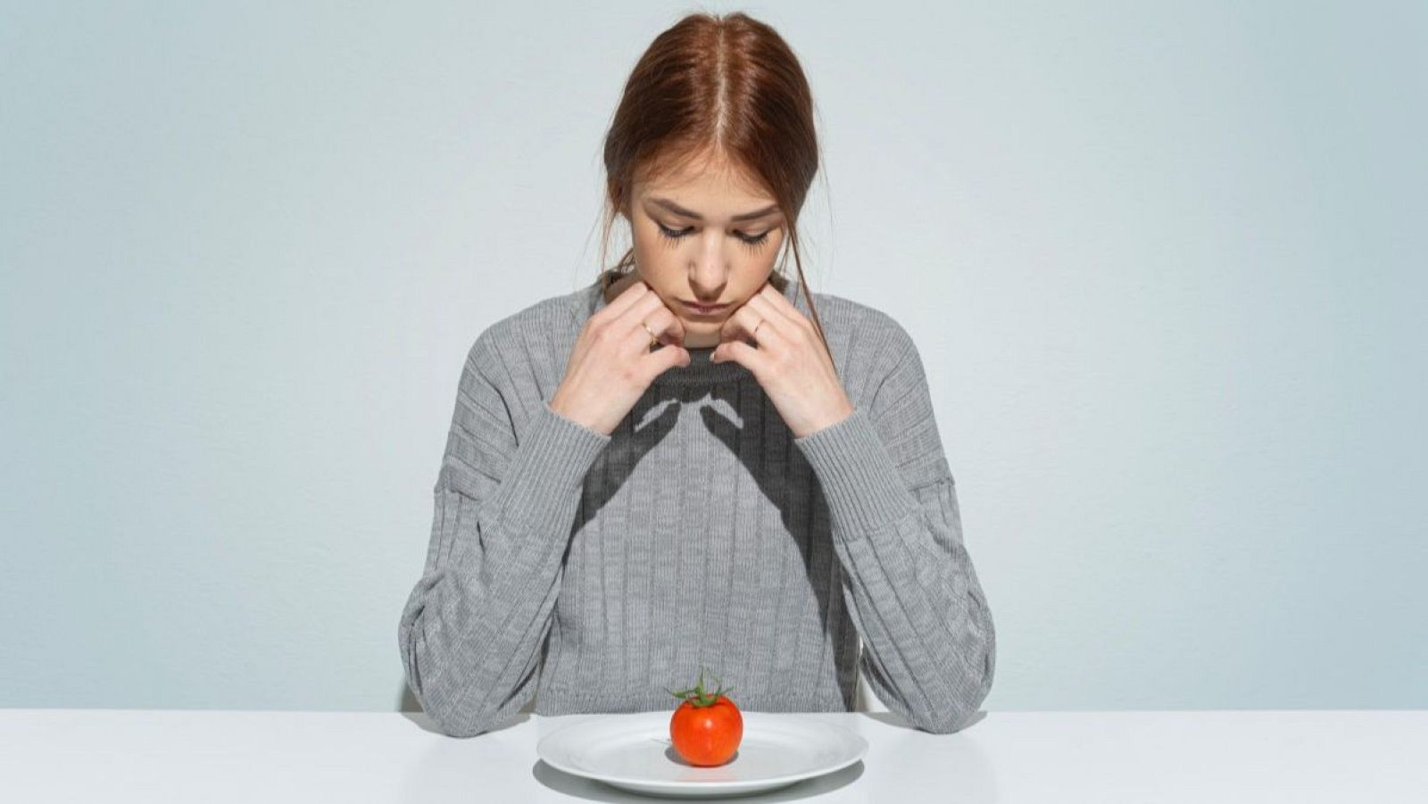 Joven con jersey gris mira un tomate en un plato, representando la anorexia y la restricción de alimentos.