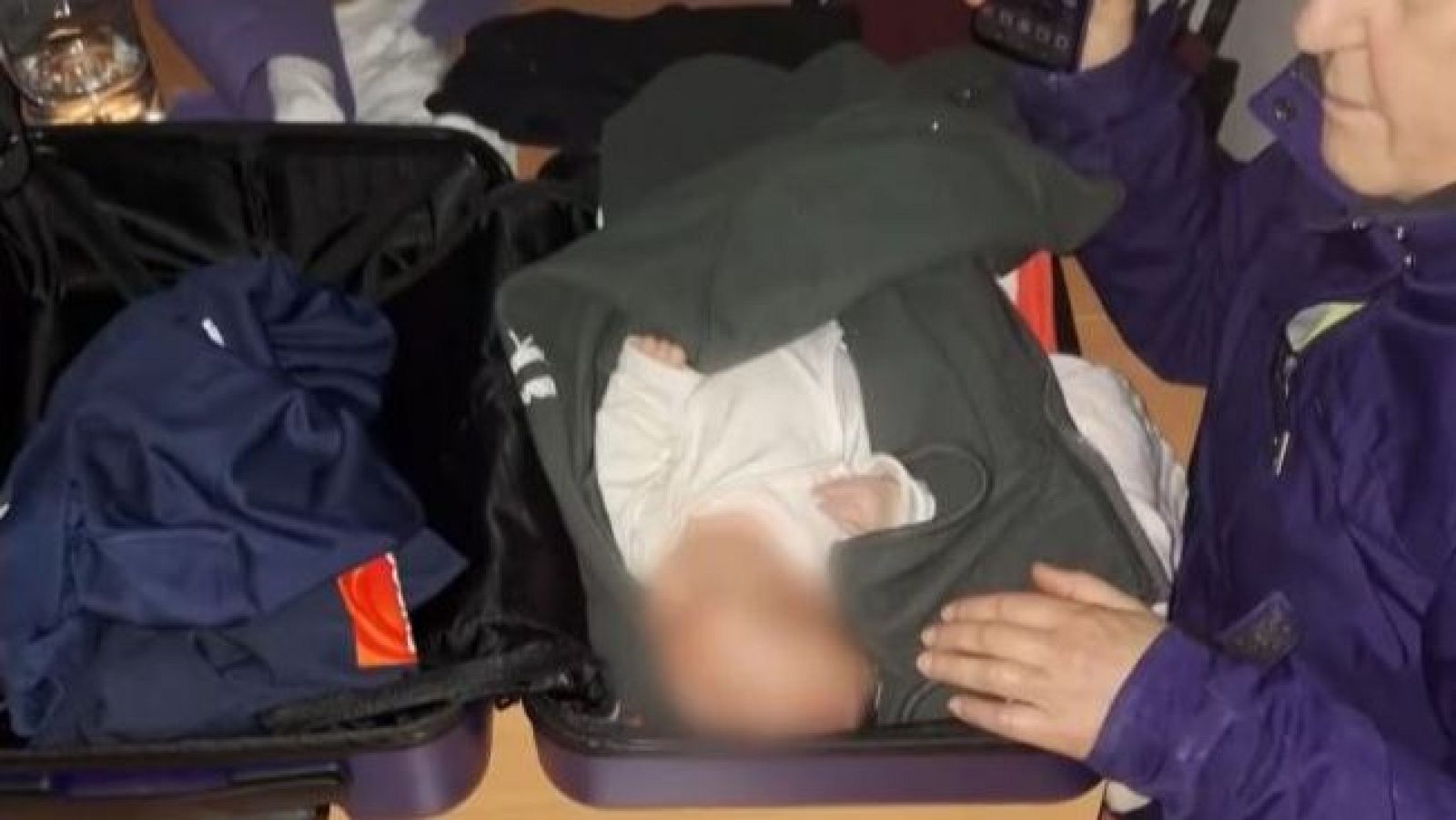 Bebé rescatado, envuelto en una sudadera gris, dentro de una maleta abierta. Una mano lo sostiene; otra maleta contiene ropa.