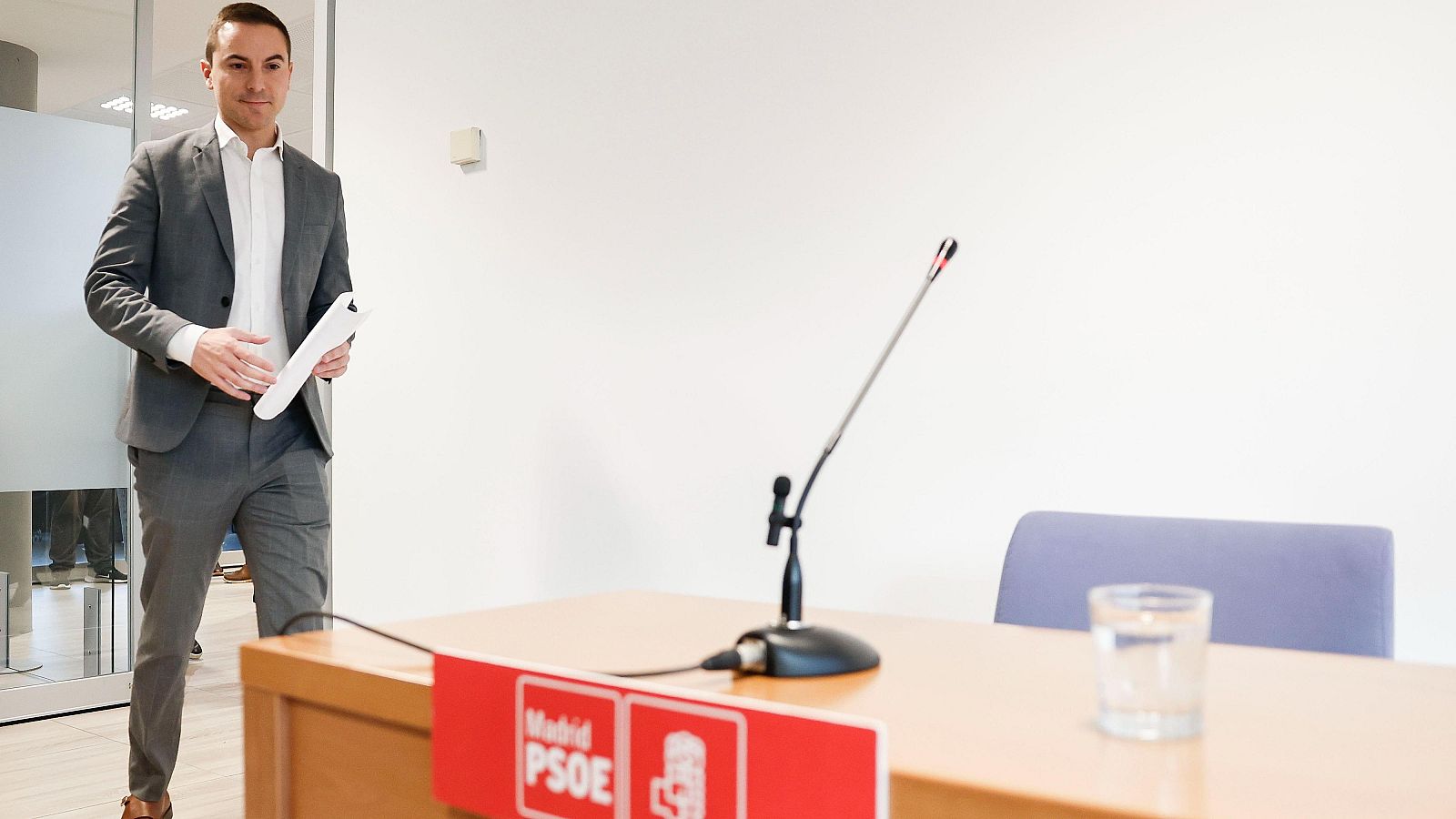 Juan Lobato (PSOE-M) llega a una comparecencia ante los medios en la Asamblea de Madrid. Lleva traje gris y papeles.