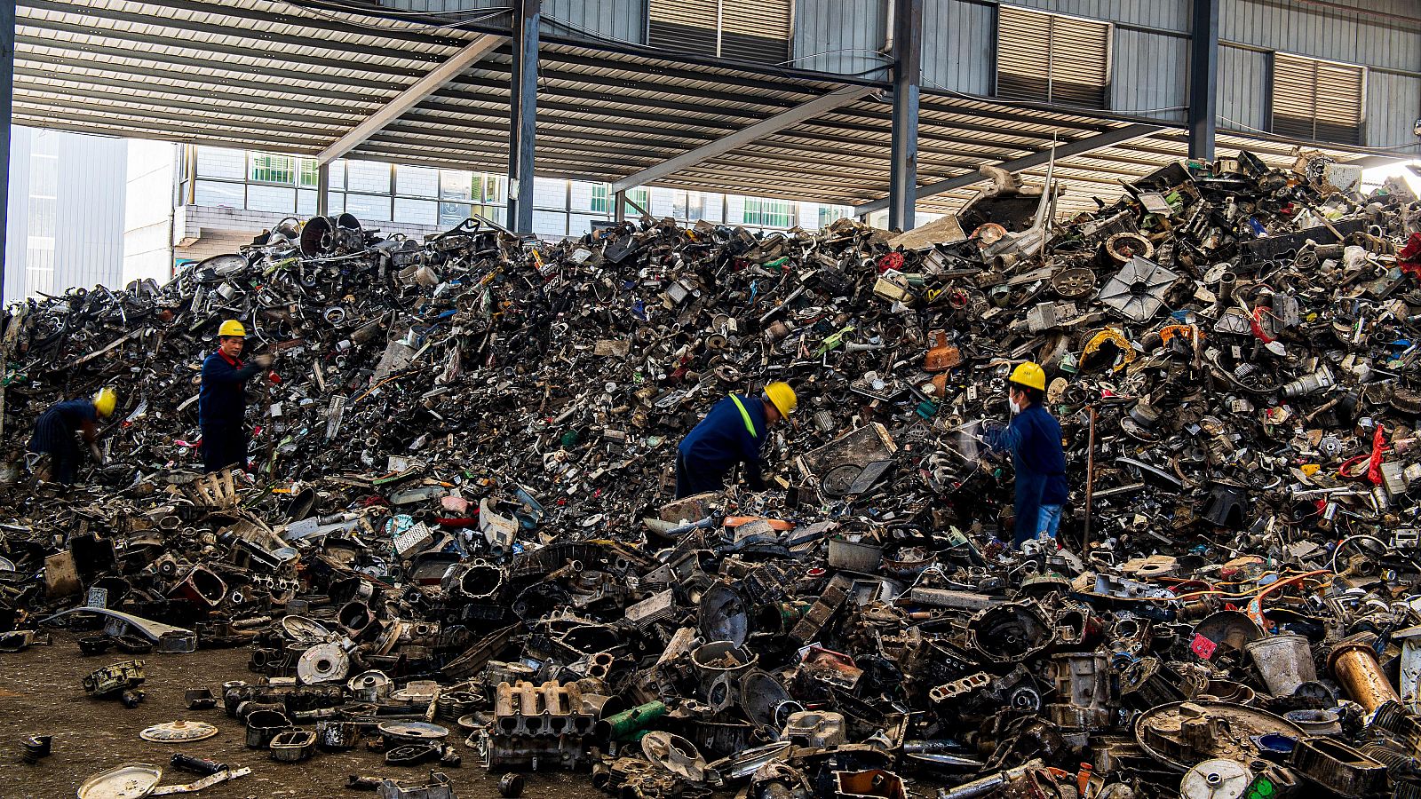 Trabajadores revisan chatarra metálica en una nave industrial.  Se ven piezas de coches y maquinaria. Reciclaje de metales.