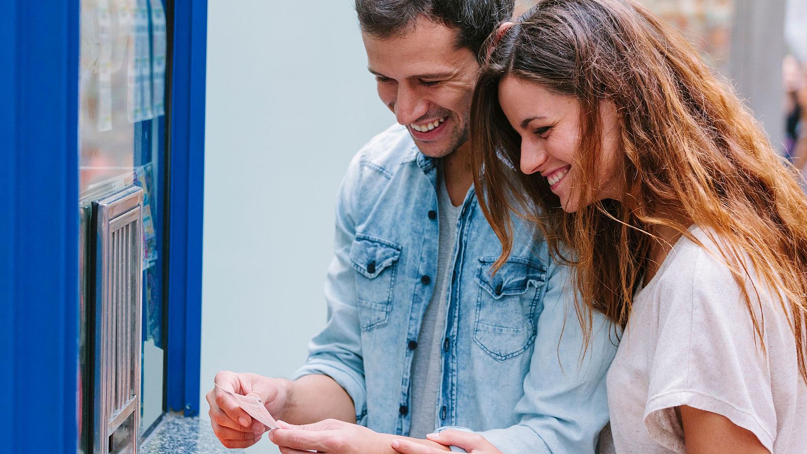 Pareja joven sonríe emocionada mirando un décimo de lotería. Él, con camisa vaquera, lo sostiene; ella, con camiseta blanca, se inclina hacia él.  Ante una máquina expendedora azul.