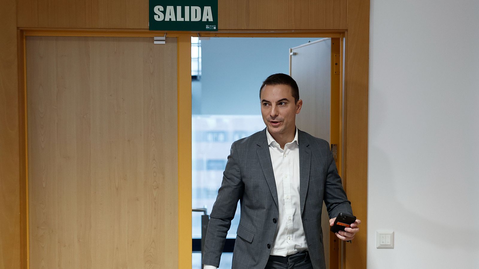 Juan Lobato (PSOE) sale por una puerta señalizada 'SALIDA', con traje gris, camisa blanca y móvil en mano.