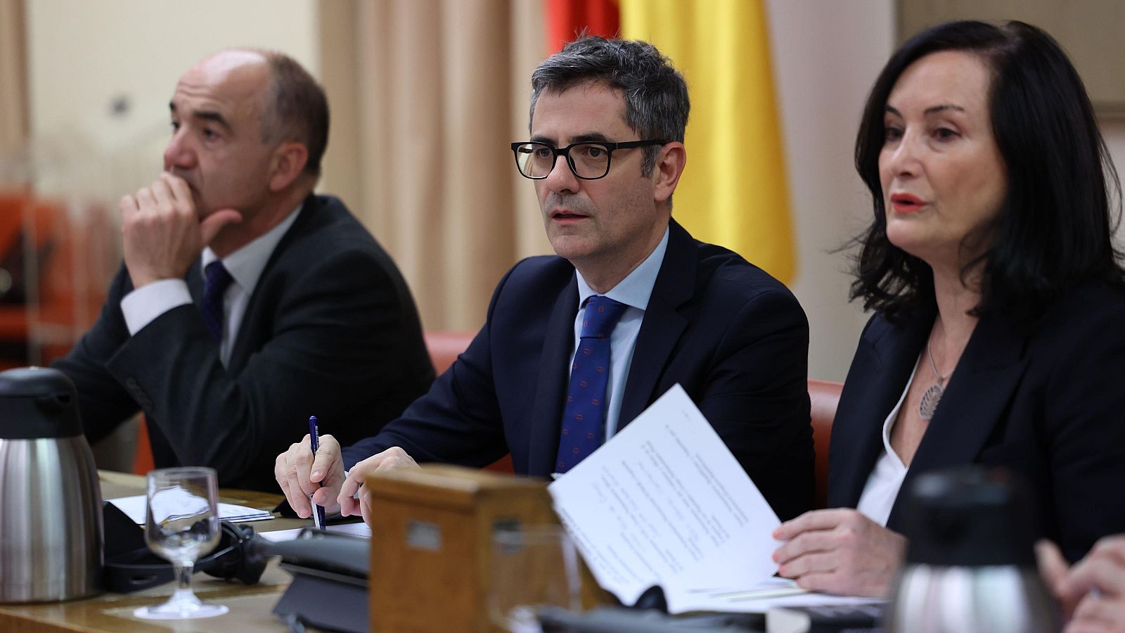 Reunión en España con el ministro Félix Bolaños y otras dos personas en traje, revisando documentos.  Bandera española visible.