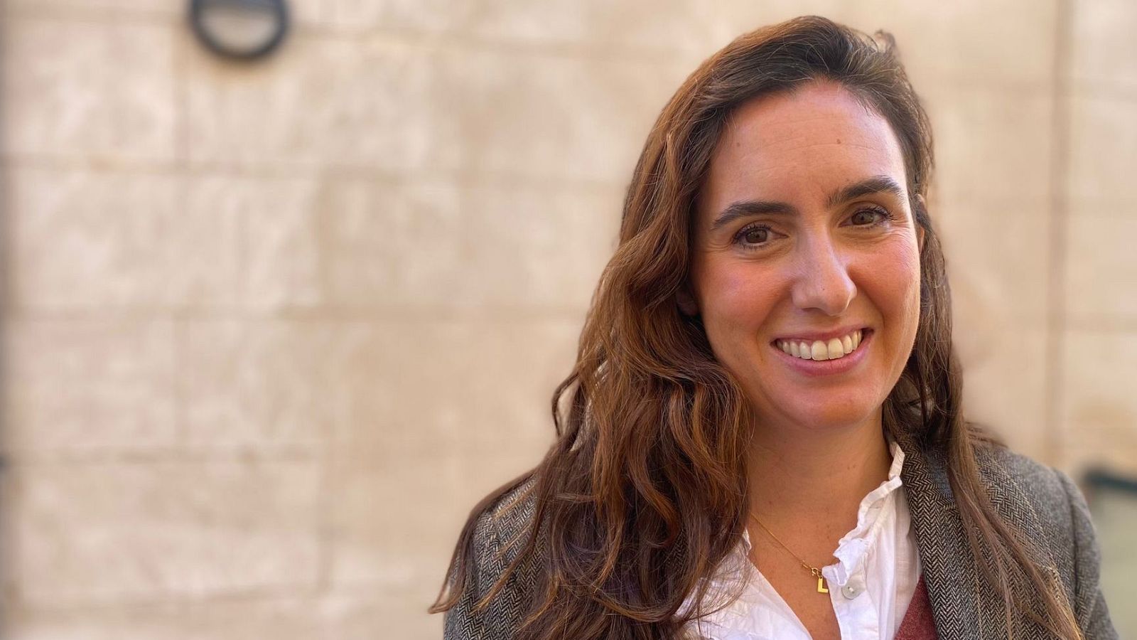 Retrato de mujer con el pelo castaño ondulado, blusa blanca, jersey granate y chaqueta gris. Sonríe y lleva un collar con colgante.