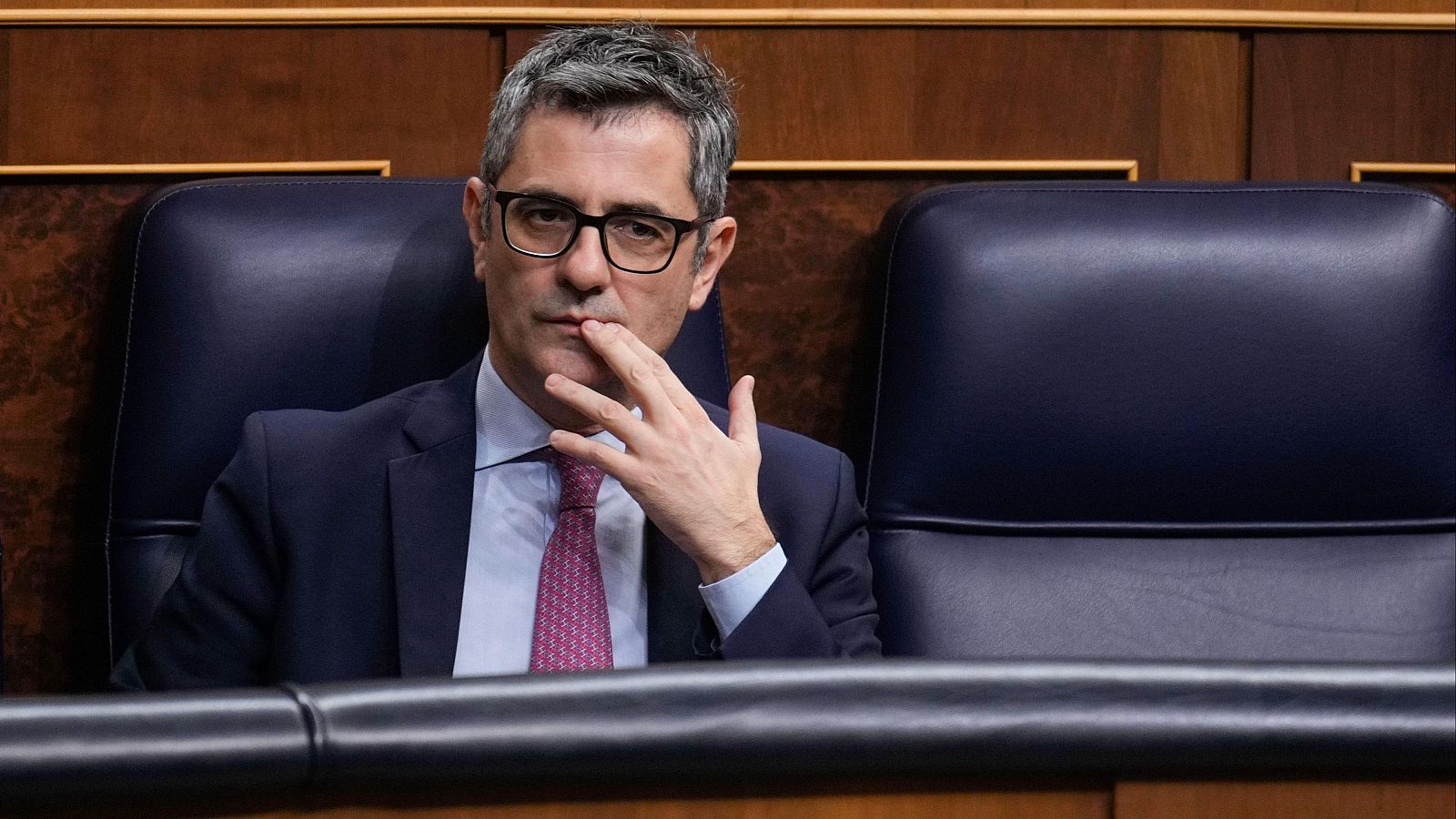 Ministro en pleno del Congreso, sentado en su escaño con traje oscuro, camisa clara y corbata rosa, un dedo sobre la boca, en actitud reflexiva.
