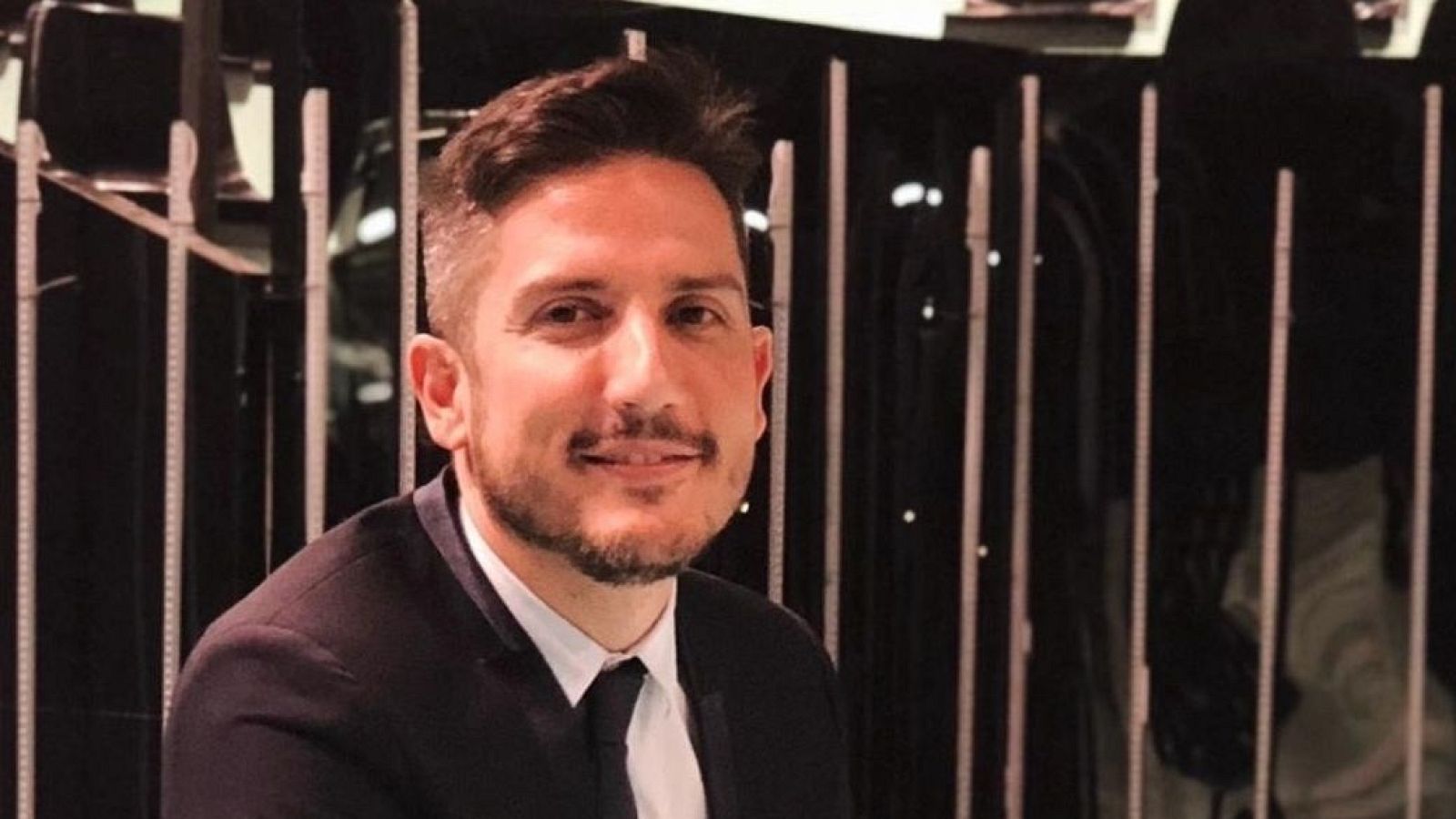 Hombre de entre 30 y 40 años con traje oscuro, camisa blanca y corbata negra, sonríe a cámara frente a un fondo metálico con barras verticales.