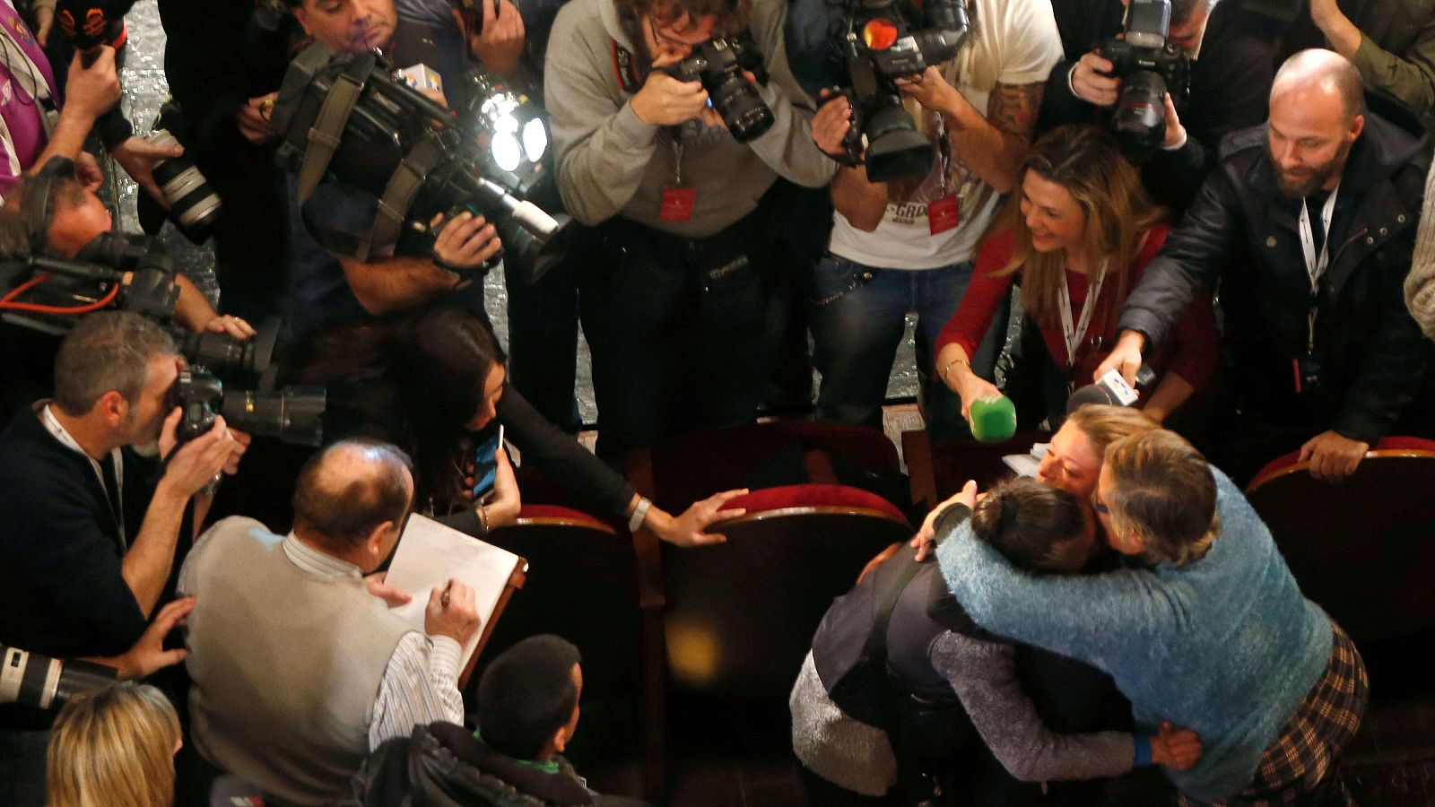 Emotivo abrazo entre dos mujeres rodeado de periodistas con cámaras y micrófonos en un espacio público con asientos rojos.  Escena de gran expectación mediática.