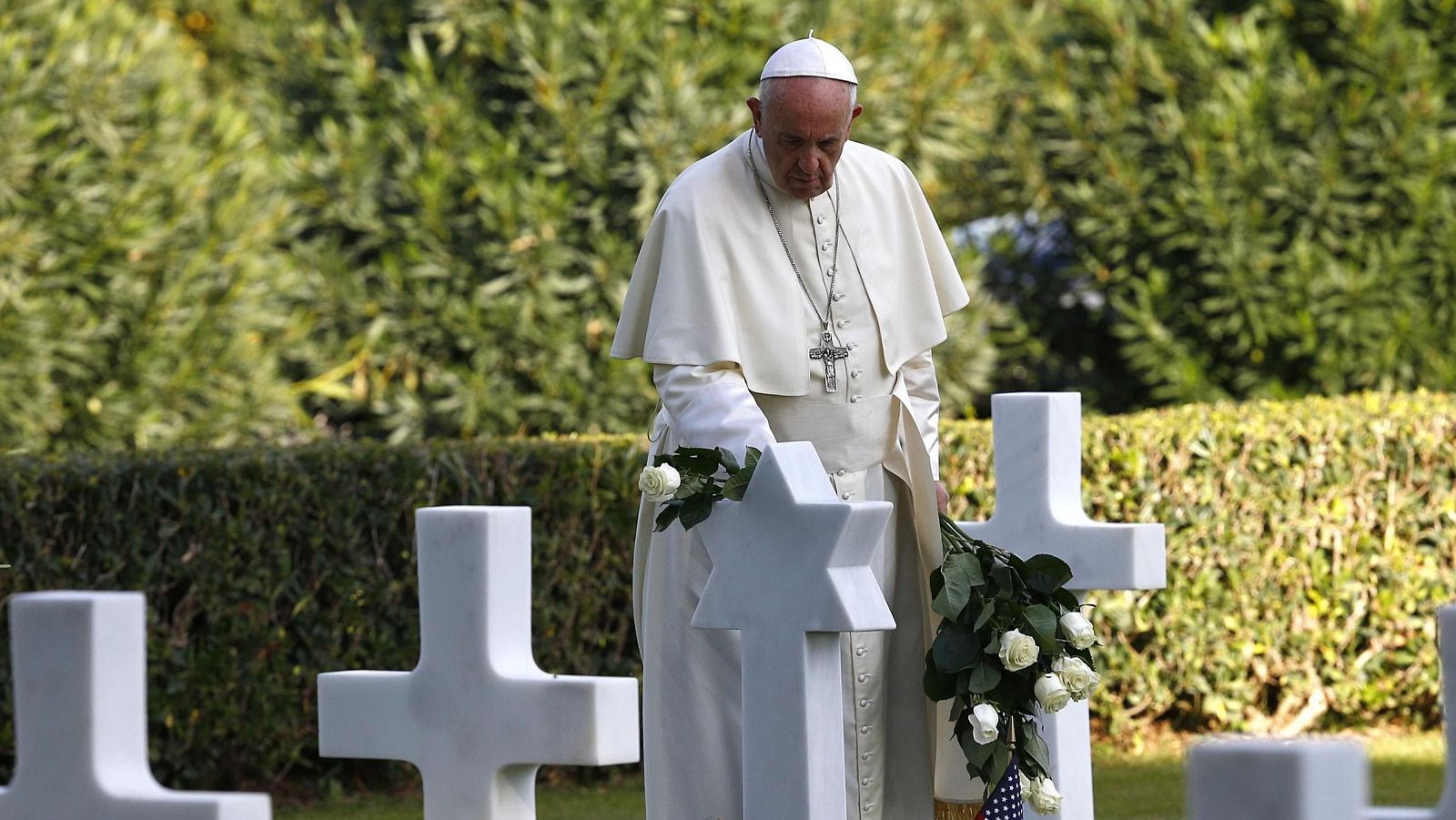 El Papa Francisco rinde homenaje en un cementerio, colocando rosas blancas sobre una lápida con estrella de David.  Se observan numerosas cruces en el entorno.
